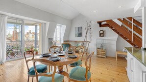 A dining area with a round table set for six, surrounded by wooden chairs with green cushions. A staircase with wooden railings leads to an upper floor.