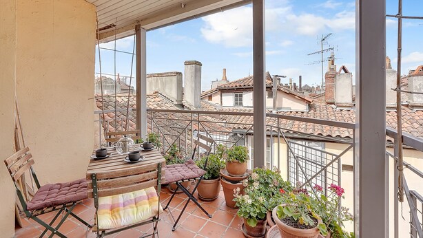 A balcony with a wooden table, chairs, potted plants, and a view of neighboring buildings.