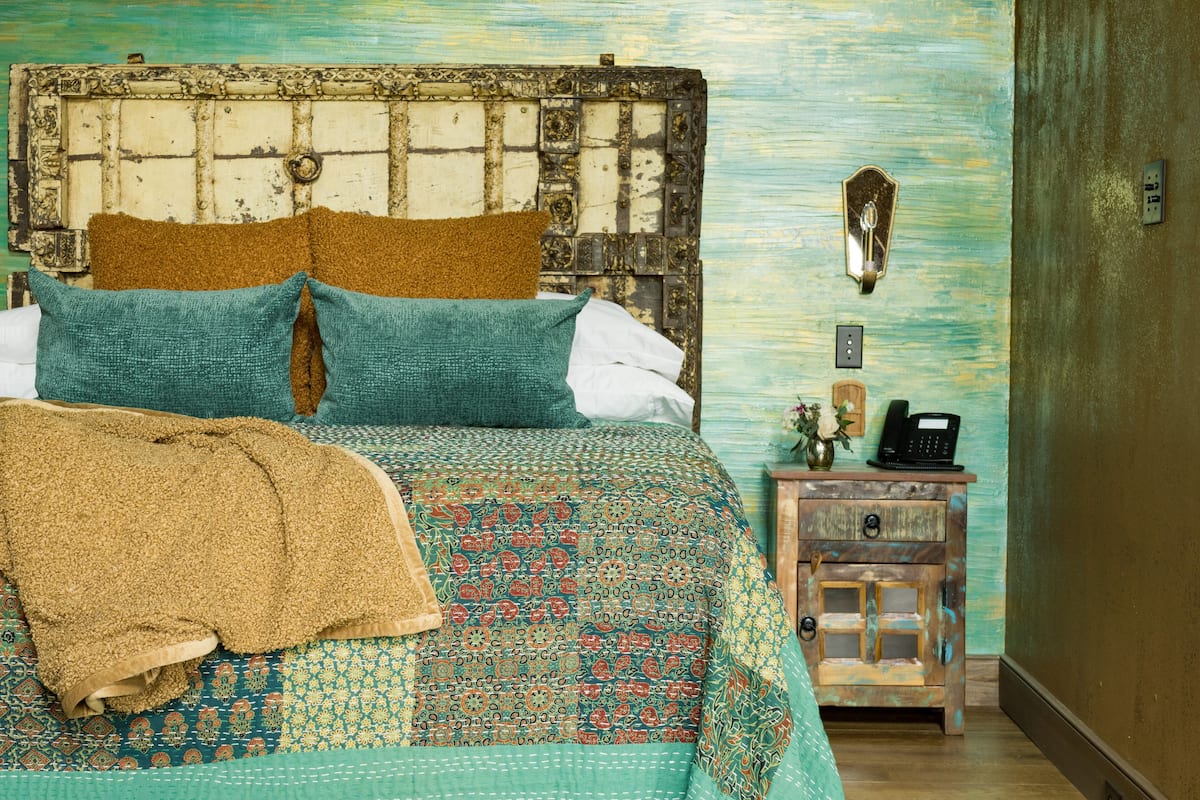 A bed with a patterned bedspread, two pillows, and a blanket. There is a wooden nightstand with a telephone and a small vase with flowers.