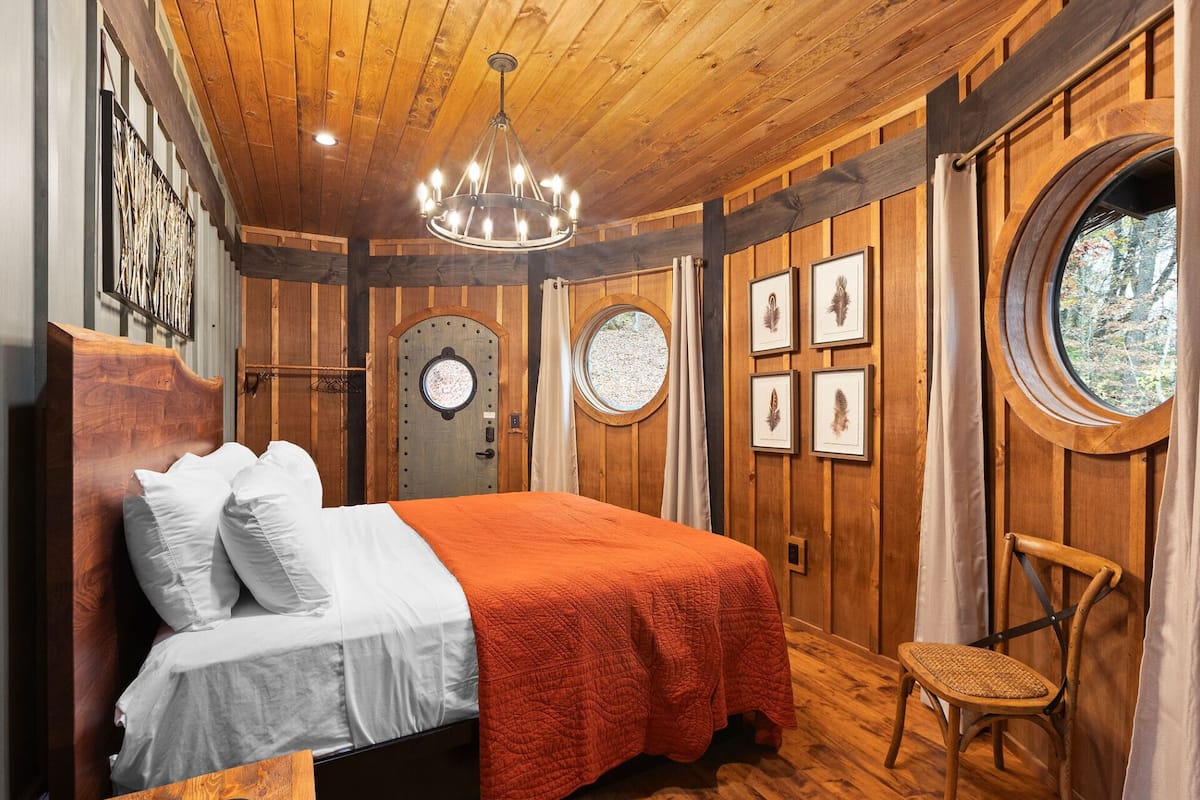 A wooden-paneled bedroom with a bed, a chandelier, a round window, and framed pictures on the wall.