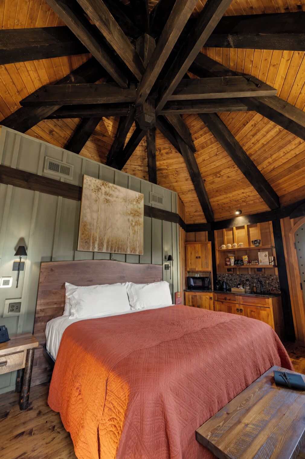 Une chambre à coucher rustique avec un plafond en bois, un grand lit recouvert d’une couverture rouge, une table de chevet en bois, une table en bois et un coin cuisine doté de placards en bois.
