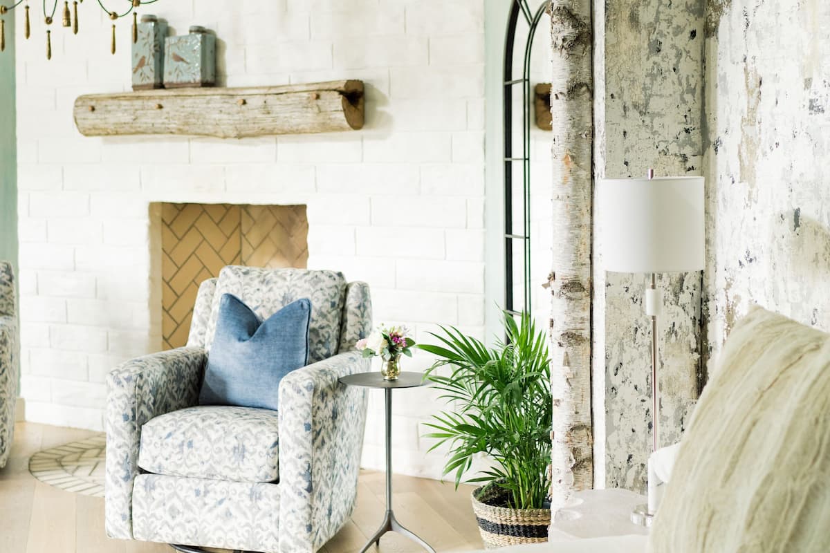 A modern bedroom with a white brick fireplace, a patterned armchair, a bed with a white headboard, a floor lamp, and a chandelier.