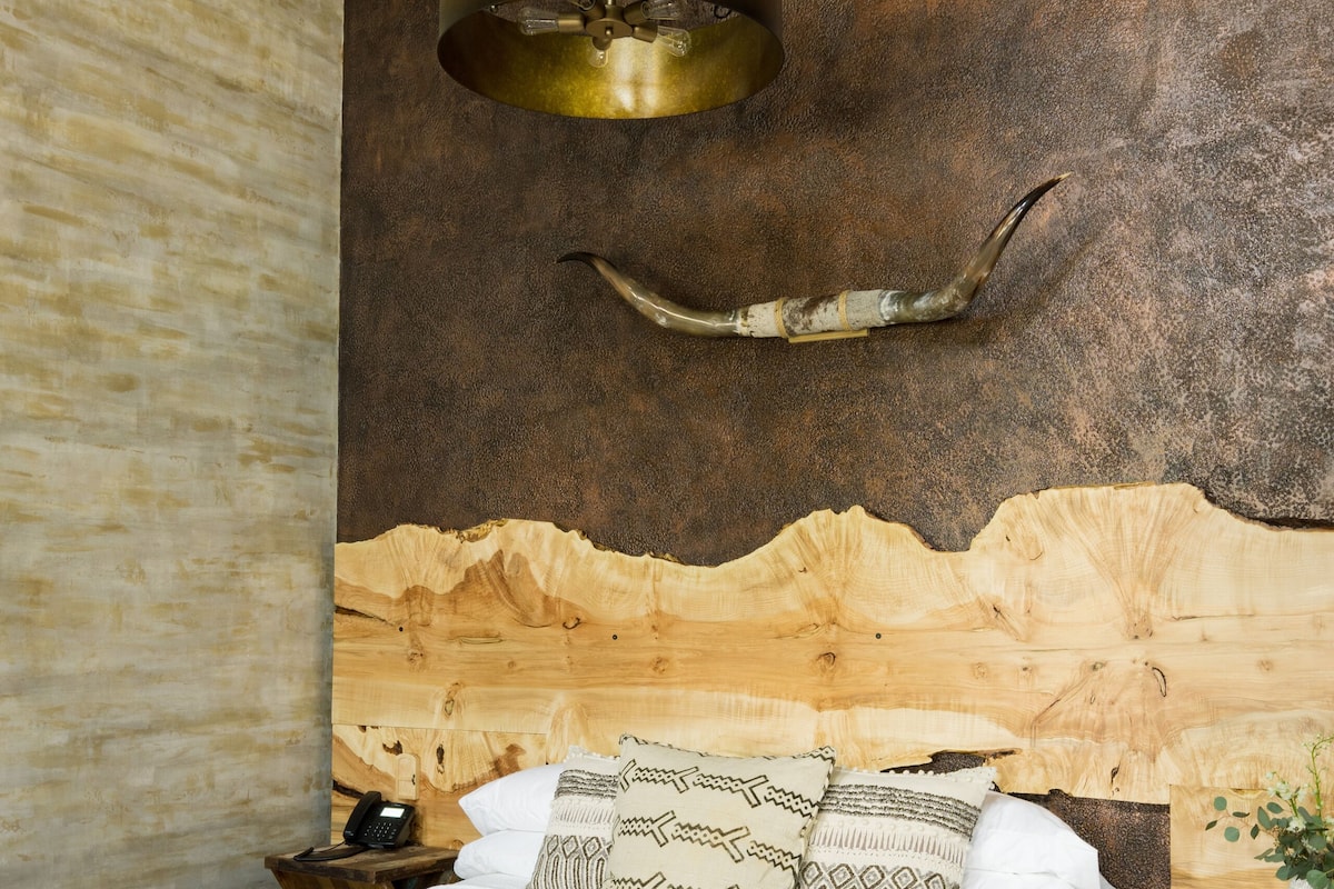 A rustic bedroom with a wooden headboard, a bed with patterned bedding, two stools with leather cushions, and a brass pendant light.