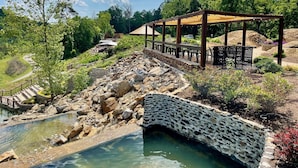 A stone bridge over a clear water body, a wooden pergola with seating, and a pathway leading to a building in a landscaped area. - Ancient Lore Village (Knoxville)