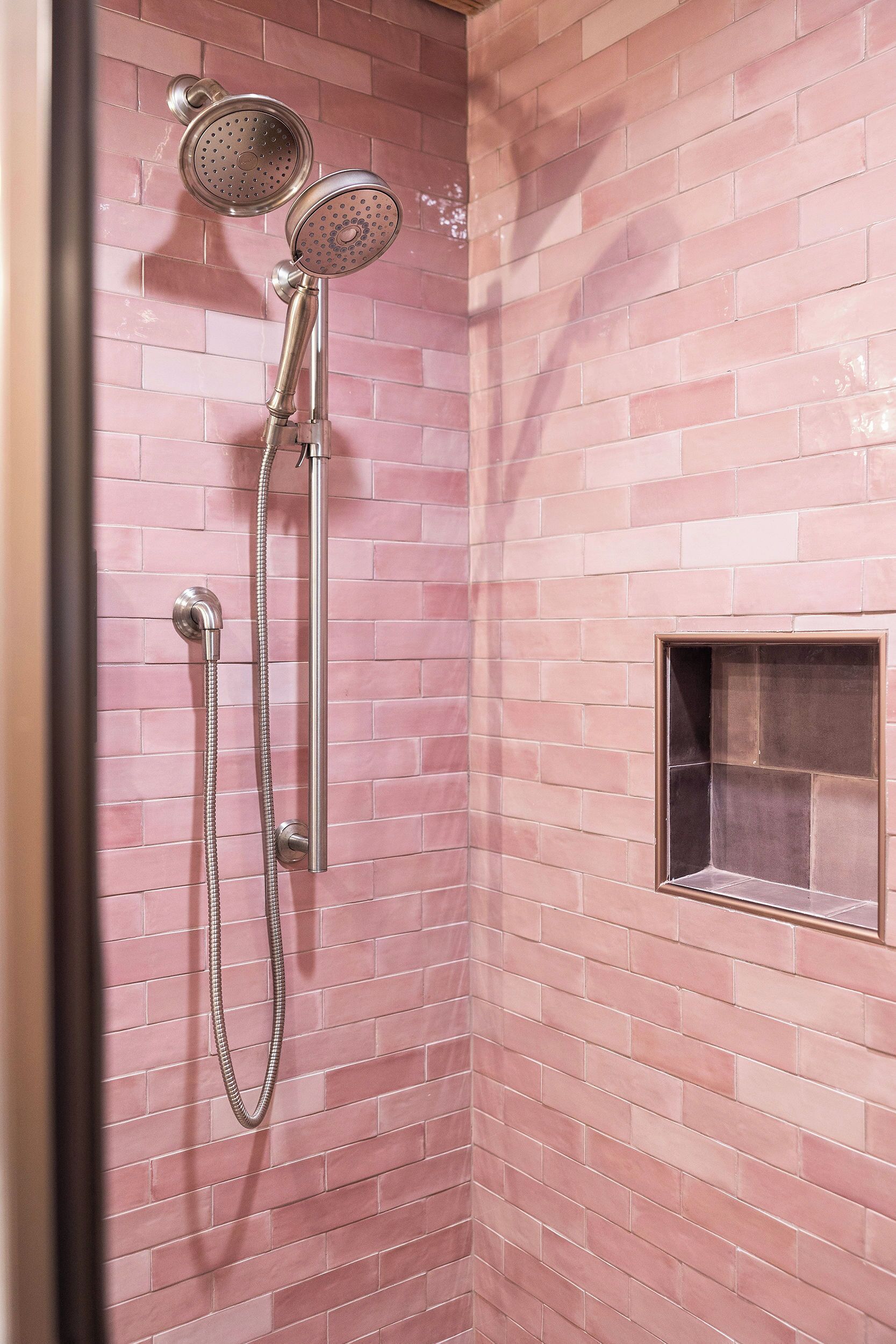 A shower with a handheld showerhead and a handheld hose, set against a pink-tiled wall.