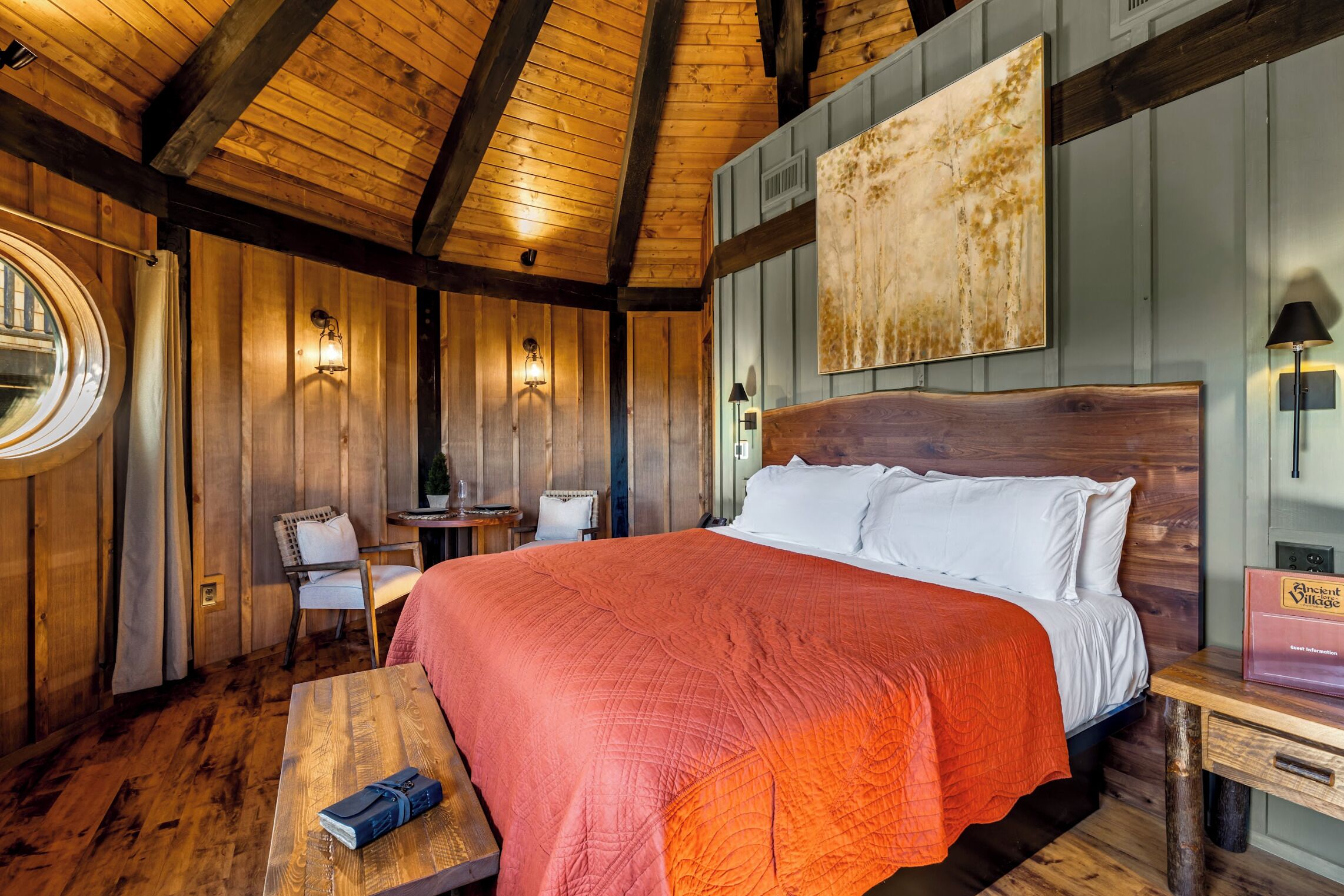 A wooden room with a large bed, a round window, and a table with a lamp.