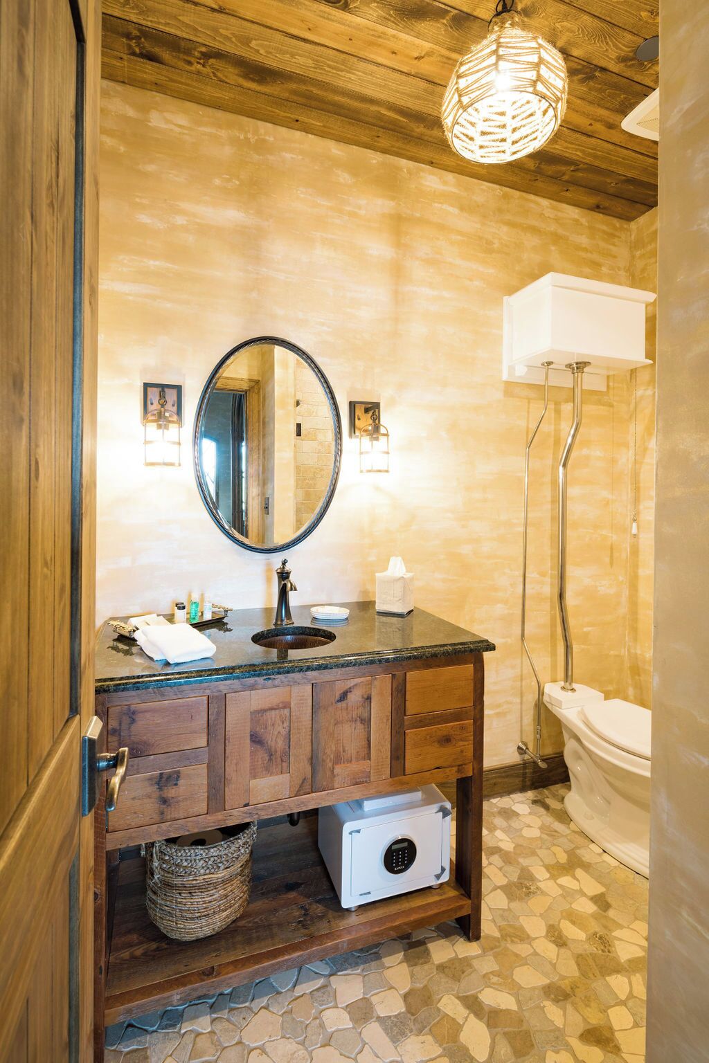 Een badkamer met een houten wastafelmeubel, een ronde spiegel, een wastafel met toiletartikelen, een wit toilet en een mozaïekvloer.