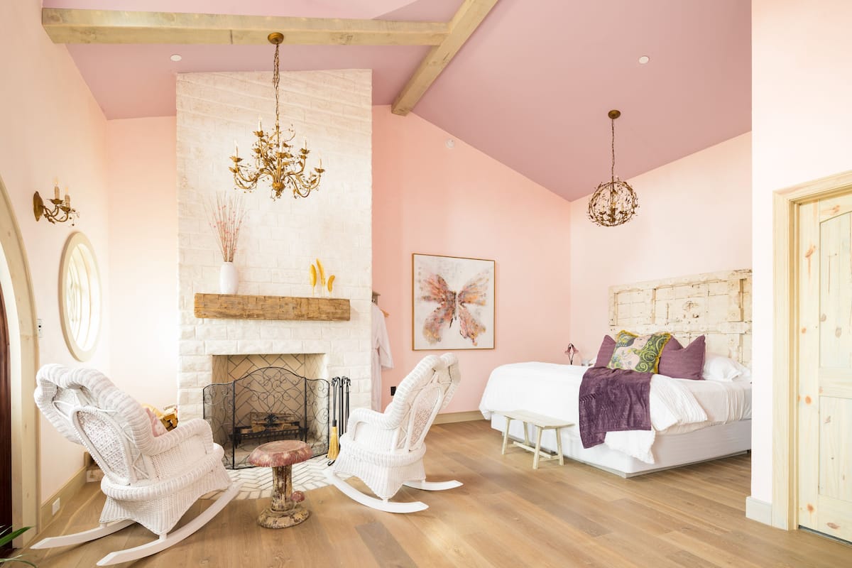 A bedroom with a white bed, a white rocking chair, a fireplace, and a chandelier.