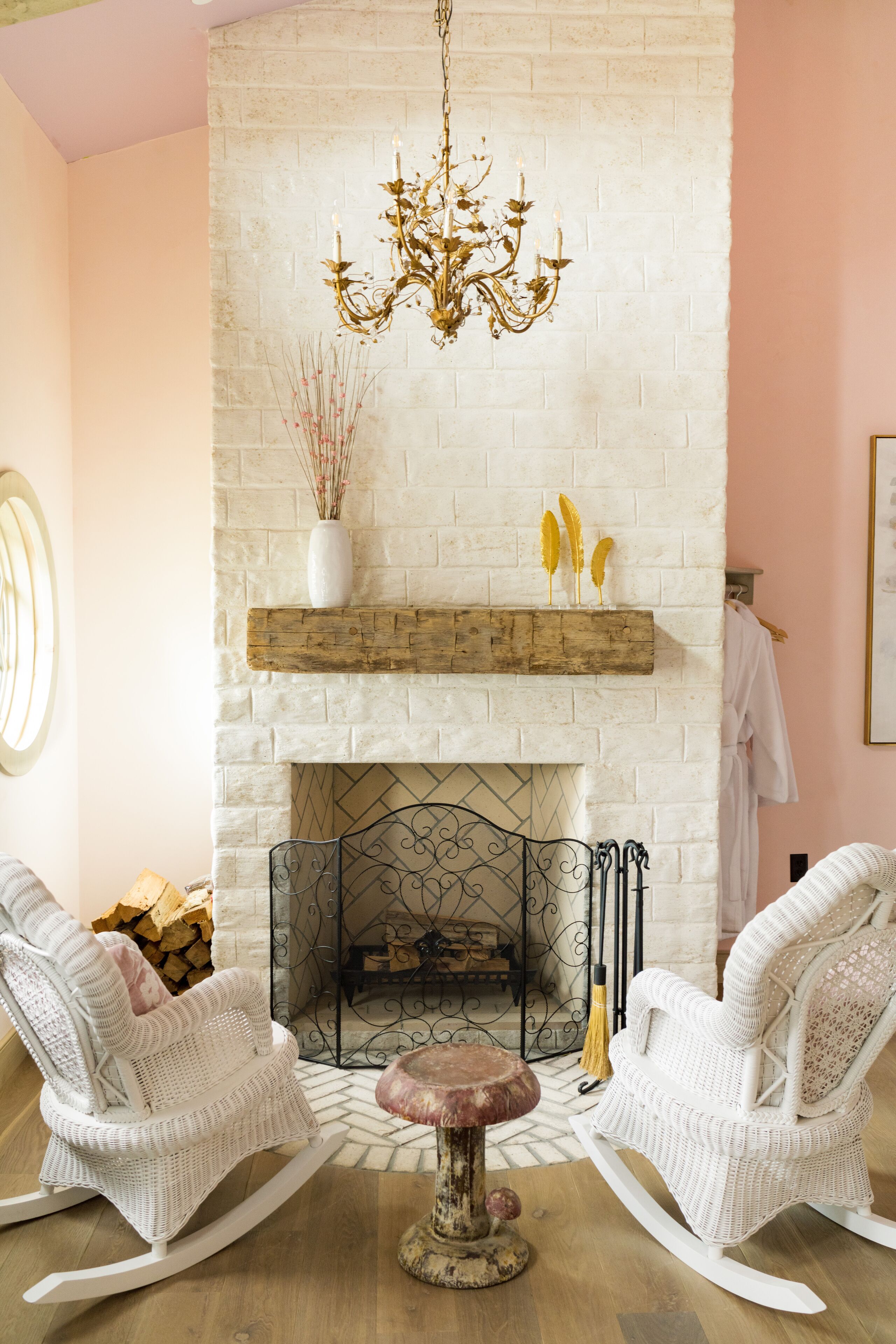 Une pièce chaleureuse avec une cheminée en briques blanches, deux fauteuils à bascule blancs et un lustre.