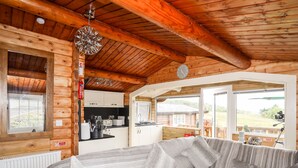 A cozy log cabin interior with a sofa, kitchen, and large windows offering a view of the outdoors. - Ty Coed - 63 (Blaenau Ffestiniog)