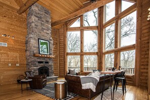 A log cabin interior with a stone fireplace, wooden walls, large windows, a leather sofa, and a dining area. - Luxury 6-Bedroom Lodge Dubuque, Field of Dreams & Galena (Durango)