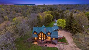A large log cabin with multiple windows and a covered porch, surrounded by a forested landscape. - Luxury 6-Bedroom Lodge Dubuque, Field of Dreams & Galena (Durango)