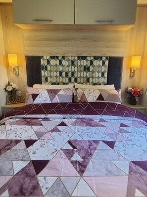 A hotel room with a large bed, a geometric-patterned headboard, two bedside lamps, and a nightstand with a vase of flowers. - 3 Bedroom Static Caravan, Hoburne Devon Bay (Paignton)