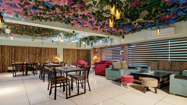 A spacious indoor area with a dining section featuring wooden tables and chairs, a seating area with sofas and ottomans, and a ceiling adorned with hanging plants and lights. - Treebo Nearmi Bell House (Gurugram)