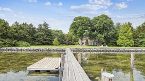 Een houten steiger die uitsteekt in een rustig wateroppervlak, met een huis op de achtergrond, omringd door bomen.