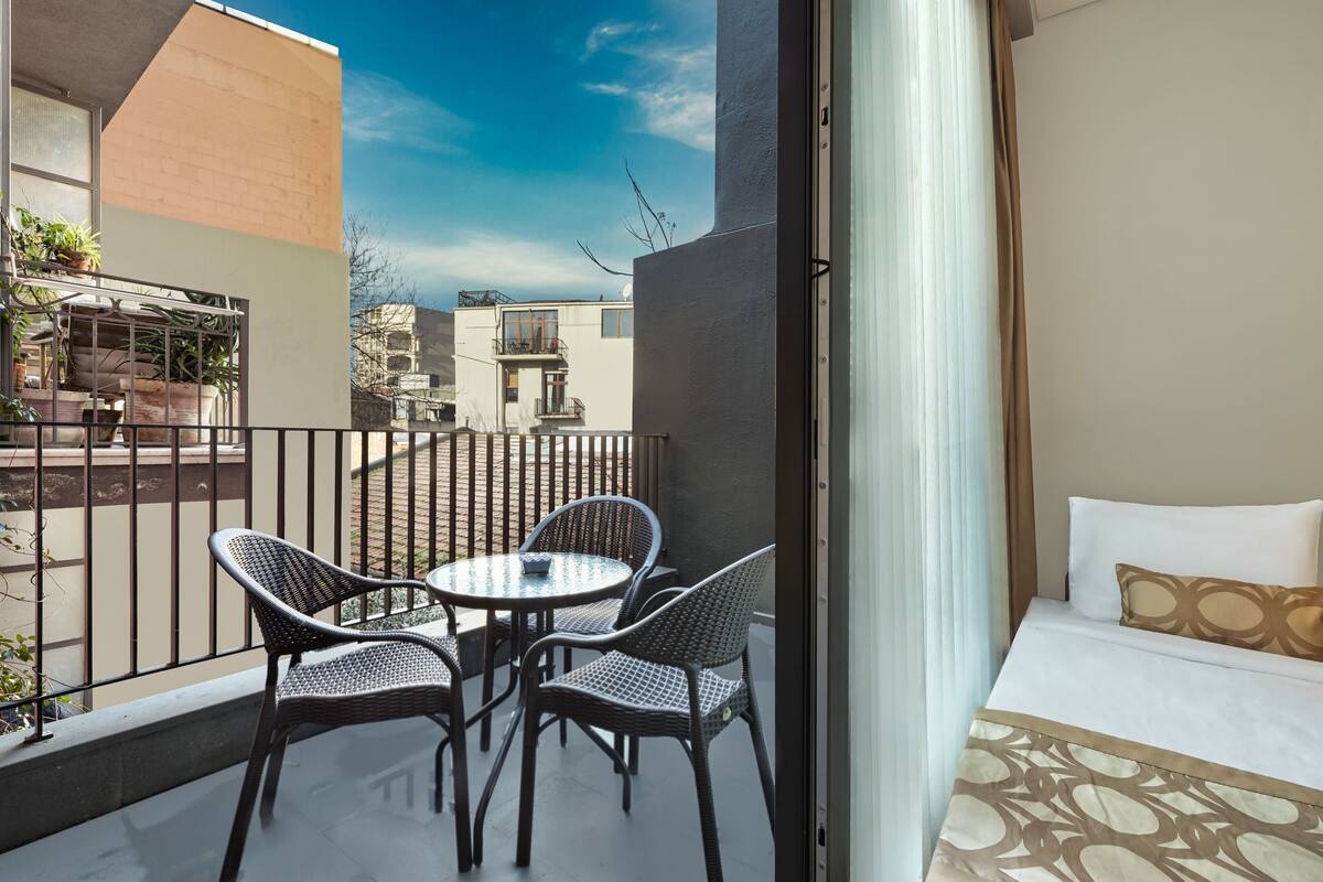 Un balcón con sillas de mimbre y una mesita, una cama con ropa de cama estampada, y vistas a edificios cercanos.
