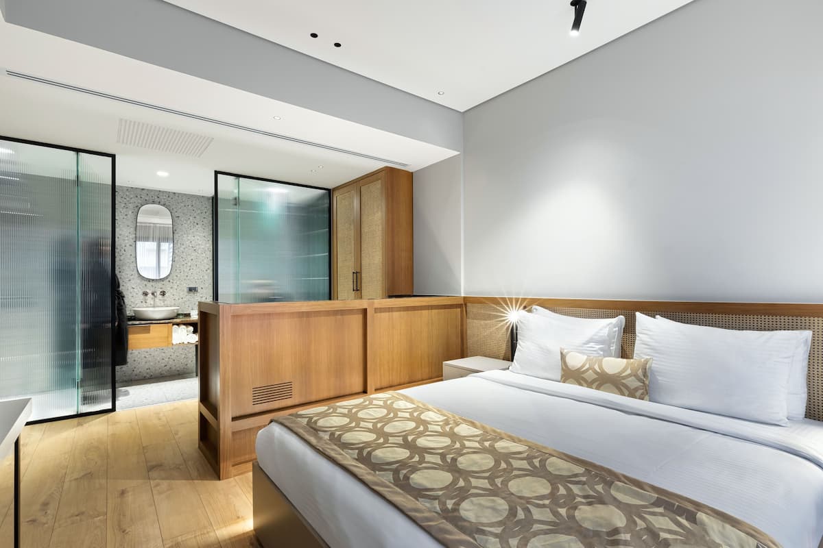 Habitación de hotel moderna con una cama grande, un armario de madera, cabina de ducha de vidrio y baño con un espejo grande y lavamanos.