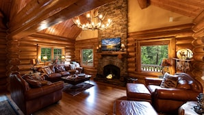 A log cabin interior with a stone fireplace, leather furniture, and a large window overlooking the forest. - Luxury Log Cabin Home at Bass Lake with Air Conditioning, with backup generator  (Bass Lake)