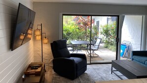 A modern living room with a black armchair, a coffee table, and a TV mounted on the wall. There is a sliding glass door leading to an outdoor patio with a table and chairs.