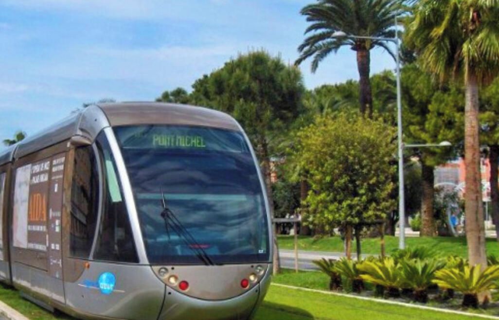 A modern bus with 'PUUTT NICHOL' on the front, parked on a road with greenery and palm trees in the background.