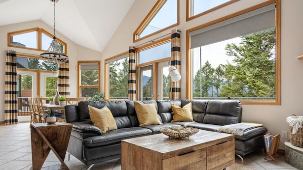 A spacious living room with a large black leather sectional sofa, wooden coffee table, and large windows offering a view of greenery outside. - Chateau Lawrence | Pool Table | Kids` Gaming Room (Fairmont Hot Springs)