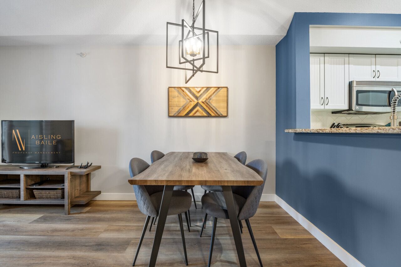 A modern kitchen with white cabinets and a blue accent wall, a dining area with a wooden table and gray chairs, and a TV on a wooden stand.