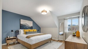 A bedroom with a bed, bedside table, lamp, and a view of the outside through a window. - High Noon | Top Floor | Pool | Hot Tub | Gym (Invermere)