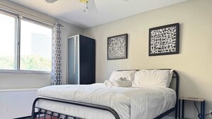 Un lit bien fait, avec du linge de lit blanc, un cadre en métal foncé et une serviette blanche posée sur les oreillers. Deux œuvres d’art abstraites ornent le mur, et une fenêtre est agrémentée d’un rideau à motifs.
