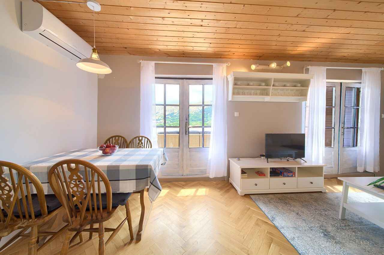 Ein gemütliches Zimmer mit Holzbalkendecke, einem Esstisch mit Stühlen, einem Fernseher auf einem weißen Schrank und großen Fenstern mit Vorhängen.