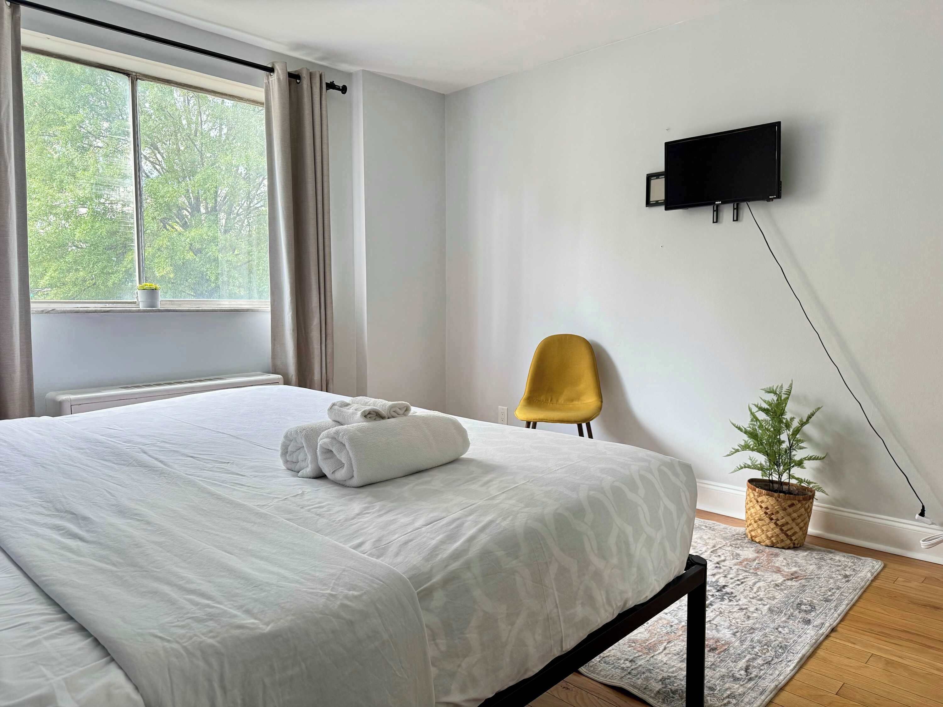A neatly made bed with white linens and rolled towels, a yellow chair, a television mounted on the wall, and a small potted plant on the windowsill.