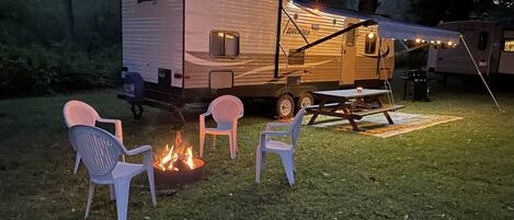 A recreational vehicle with a picnic table and chairs, a fire pit, and a tent set up in a grassy area.