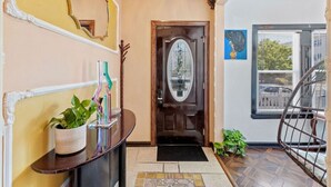 Un corridoio con una porta in legno, uno specchio, una pianta in vaso, un tappetino nero e una sedia in vimini.