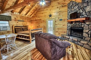 A log cabin interior with a stone fireplace, wooden ceiling beams, a bed, a couch, and a dining area.
