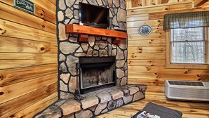Une cheminée en pierre avec un téléviseur au-dessus, une pancarte indiquant « Bienvenue au chalet », et une fenêtre donnant sur la forêt.