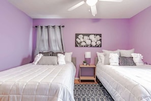 Une chambre avec deux lits, un ventilateur de plafond, un tableau représentant des fleurs accroché au mur et un tapis à motifs.