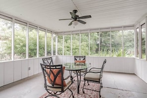 Une terrasse couverte avec une table et des chaises en verre, un ventilateur de plafond et une vue sur la verdure environnante.