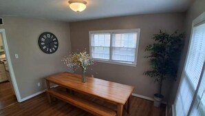 A dining area with a wooden table and bench, a wall clock, a window with blinds, and a potted plant. - Near Midtown, Highways, Downtown! Bel-Air Bungalow (Mobile)