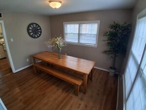 A dining area with a wooden table and bench, a wall clock, a window with blinds, and a potted plant. - Near Midtown, Highways, Downtown! Bel-Air Bungalow (Mobile)