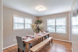 A dining area with a wooden table, chairs, and a bench. There are two windows with blinds, a ceiling light, and a wall-mounted artwork.