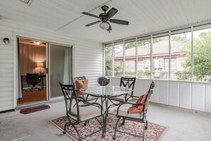 A covered patio area with a round glass table and metal chairs, a ceiling fan, and a view of a house through large windows.