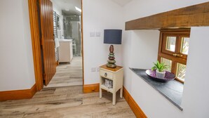 A hallway with a wooden floor, a small bedside table with a lamp, and a window with a plant. - Torties Cottage (Brampton)
