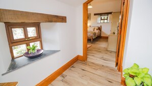 A hallway with wooden flooring, a window with a plant, and a mirror reflecting a bedroom with a bed and dresser. - Torties Cottage (Brampton)