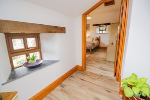 A hallway with wooden flooring, a window with a plant, and a mirror reflecting a bedroom with a bed and dresser.