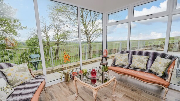 A sunlit room with a large window, a cushioned bench, a glass table, and a view of a green landscape. - Torties Cottage (Brampton)