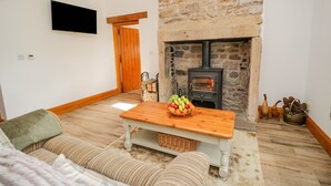 A cozy living room with a stone fireplace, a wooden table with fruit, a sofa, and a television on the wall. - Torties Cottage (Brampton)