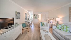 A modern living room with a flat-screen TV, a glass coffee table, a sofa, and a dining area with a table and chairs. - Renovated Studio at Naples Beach & Town! (Naples)