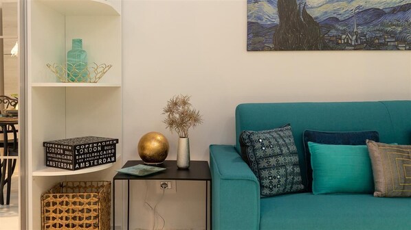 A teal sofa with patterned cushions, a small table with a vase and a globe, and a bookshelf with decorative items.