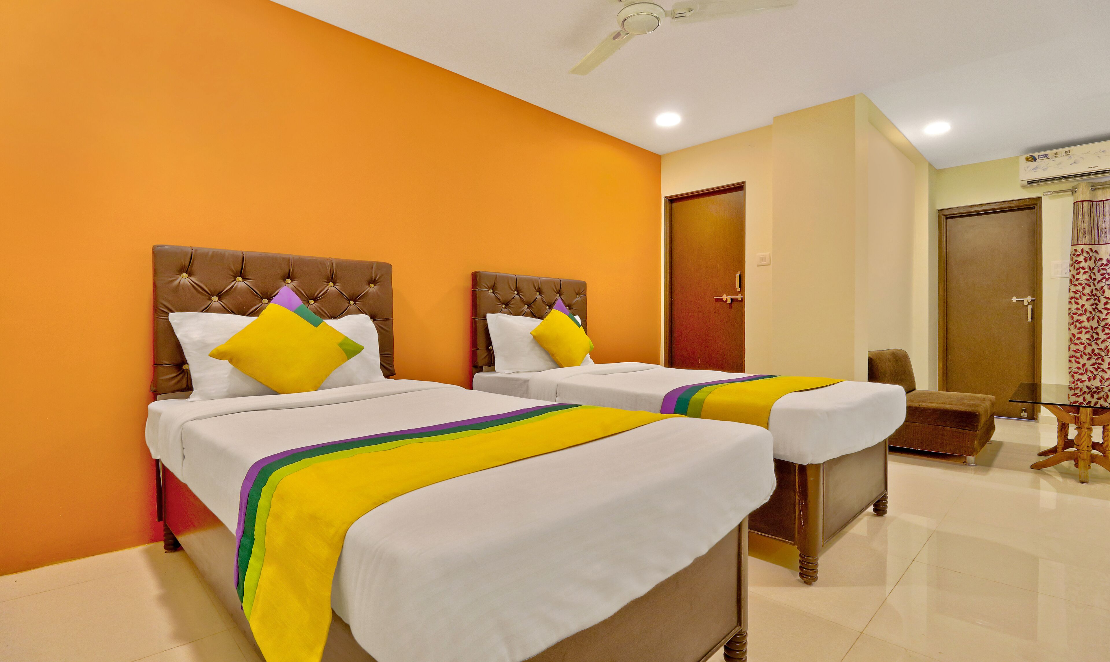 A hotel room with two beds, each with a tufted headboard, white bedding, and colorful pillows. There is a wooden chair and a small table with a glass top.