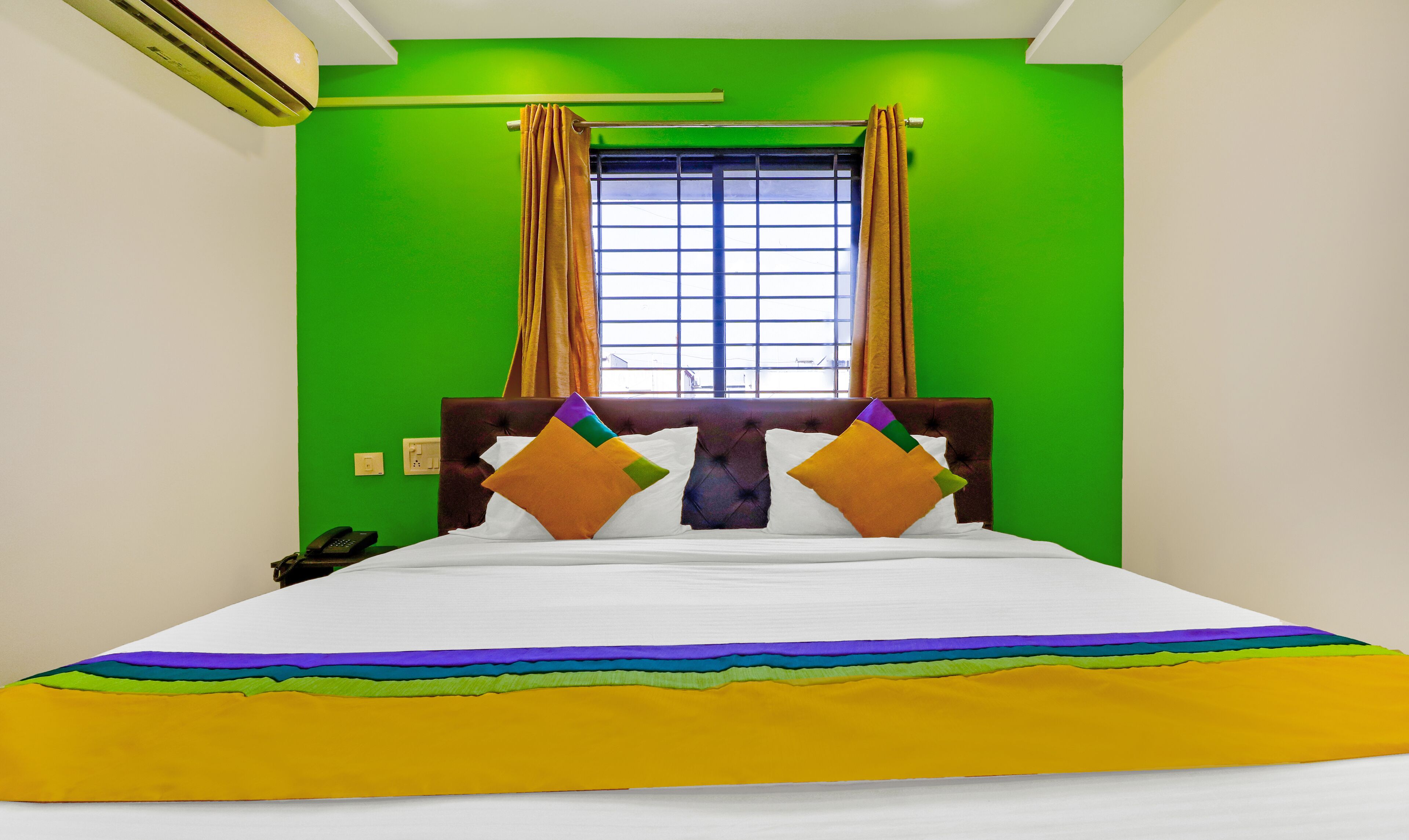 Habitación de hotel con pared verde, ventana con cortinas, cama con colchón blanco y almohadas coloridas, y teléfono junto a la cama.