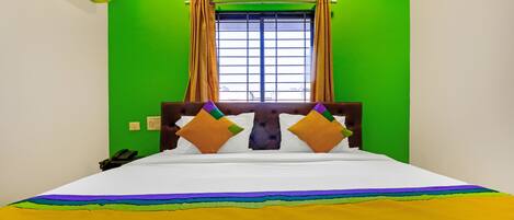 Habitación de hotel con pared verde, ventana con cortinas, cama con colchón blanco y almohadas coloridas, y teléfono junto a la cama.